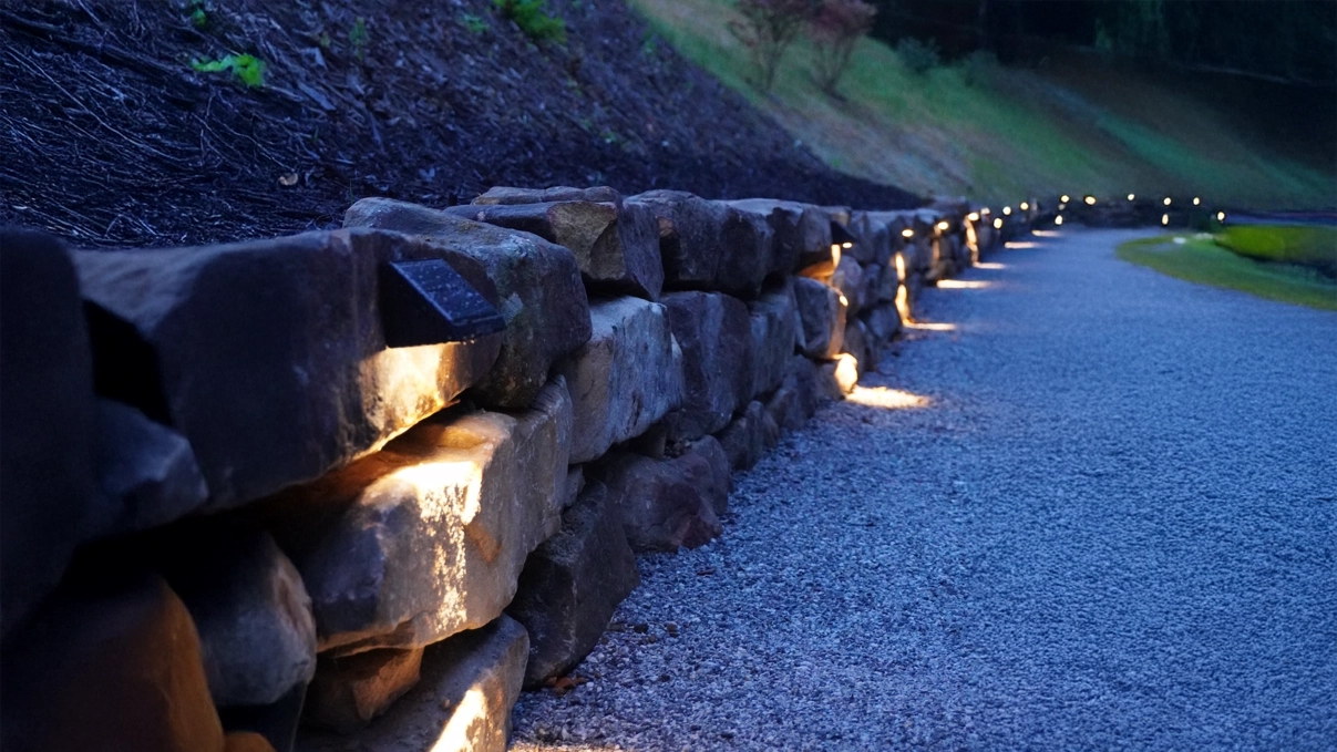 Illuminated stone retaining wall with integrated landscape lighting along a gravel pathway at dusk.