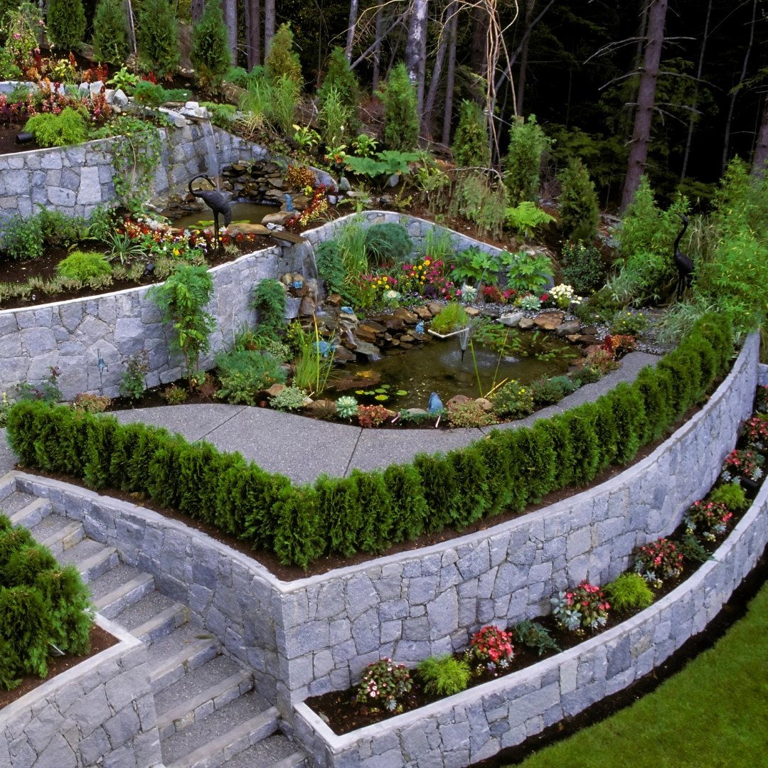 Tiered stone retaining walls with landscaped garden beds, steps, and decorative water features, showcasing advanced hardscaping and landscape design services.